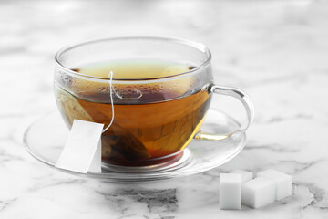 Tea bag in glass cup with hot drink and sugar cubes on light marble table, closeup