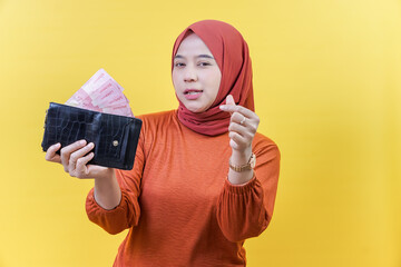 An asisan muslim woman in a hijab holds a wallet full of cash, making a hand gesture as if offering or asking for money.
