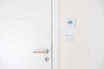 Naklejka premium Minimalistic interior with a modern white door featuring a stainless steel handle and lock. A digital thermostat displays 19.4 above a light switch. Captured from a straight-on angle. No people.