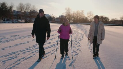Senior woman practicing Nordic walking on a frozen lake at sunset, enjoying an active winter walk with her adult son and daughter, symbolizing family and well-being. - Powered by Adobe