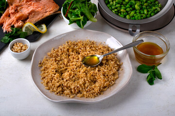 Cooked quinoa and a spoon with dressing on a ceramic plate on a white table. Around it are other...