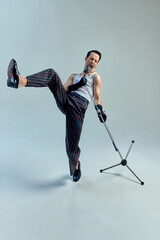 Young stylish man, singer kicking in expressive rhythm while gripping microphone, fusing dance and...