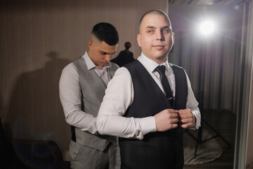 Two men are getting dressed up, one of them is adjusting his tie. The scene is set in a room with a mirror and a chair