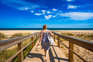 Middle aged woman tourist walking to beautiful sunny sandy beach Meia Praia on wooden promenade. Vacation in Lagos, Algarve, Portugal. Back view	