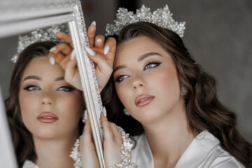 A woman is standing in front of a mirror, wearing a tiara and a white dress. She is holding a...