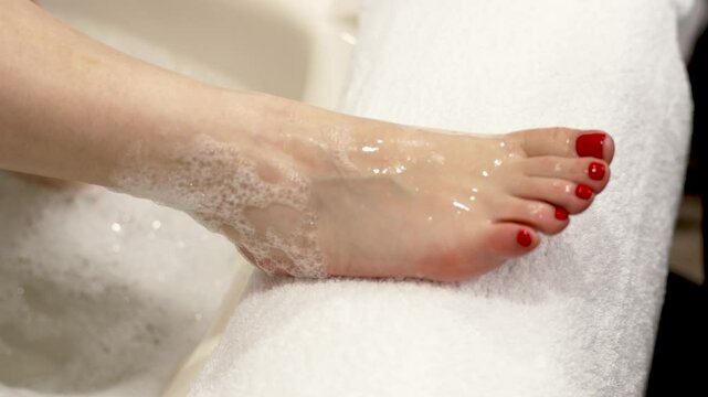 A woman takes her foot out of a bath with soapy water and places it on a pedicure stand, red gel polish pedicure, preparation for pedicure and softening the skin