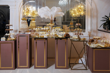 A table with a lot of food and drinks, including a lot of cupcakes. The table is decorated with gold and white items, and there are several people in the background. Scene is celebratory and festive