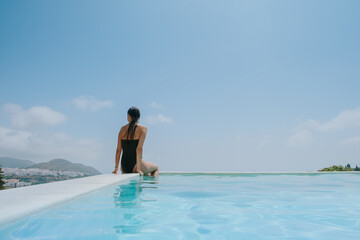A beautiful young and slender woman is sitting on the edge of a pool with long and brown hair soaked. The water is crystalline and blue and the edge of the pool joins the bright blue summer sky