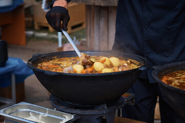 The cook stirs a thick, delicious soup in a large cast iron cauldron with a slotted spoon. National Asian food. Cooking meat dishes outdoors. Selective focus.