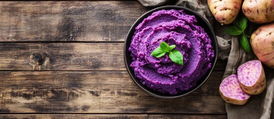 Top view of creamy purple yam mash in a bowl with fresh mint leaves beside whole purple yams on a rustic wooden table with ample copy space.