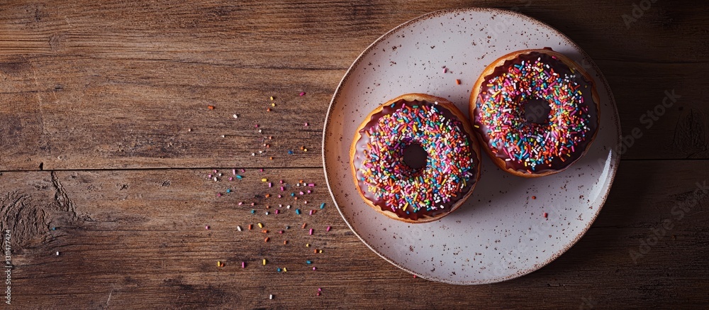 Poster Aerial view of two glazed donuts with colorful sprinkles on a round plate against a rustic wooden background with scattered sprinkles to the side