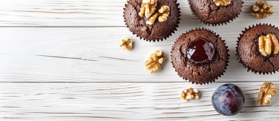 Chocolate muffins topped with walnuts and jelly plums arranged on a white wooden table with a rustic texture and natural light highlighting details.