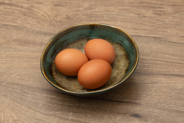 Three brown eggs are placed in a handcrafted ceramic bowl, showcasing their smooth shells against a natural wooden table. This setting adds warmth and homeliness to the kitchen environment