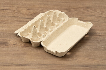 An open egg carton sits on a wooden table. It shows empty spaces where eggs are usually placed, suggesting recent cooking or meal preparation in a simple kitchen setting