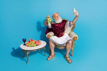Full size photo of aged man halloween emperor costume smart phone make selfie hold grape isolated on blue color background