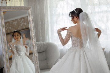 A woman in a wedding dress is standing in front of a mirror. She is wearing a veil and a necklace