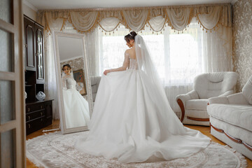 Fototapeta premium A woman in a white dress is standing in front of a mirror in a room. She is wearing a veil and a long white gown