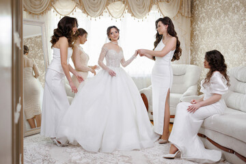 A group of women are getting ready for a wedding. One of the women is wearing a white dress and is...