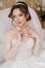 A woman in a wedding dress is wearing a necklace and a tiara. She is sitting on a couch