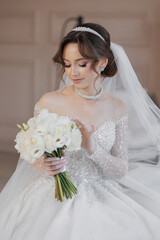 A bride is sitting on the floor with a bouquet of white flowers in her lap. She is wearing a white...