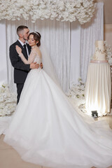 A bride and groom are posing for a picture in front of a white backdrop. The bride is wearing a long white dress and the groom is wearing a suit. Scene is happy and romantic