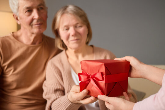 A senior couple sits together on a couch, excitedly watching as a family member presents them with a red gift box. The warm atmosphere captures a moment of joy and surprise