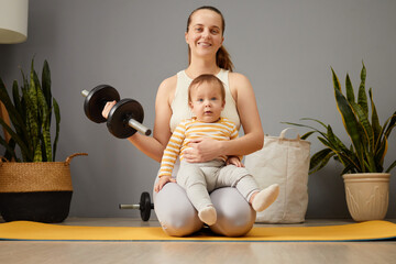 A mother sits on a yoga mat, smiling as she lifts a dumbbell in one hand while holding her baby in...
