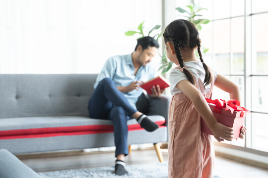 Asian cute little daughter holding and hiding wrapped gift box behind her back, making surprise for her dad, greeting young man with father's day or birthday, father sitting and reading book on sofa