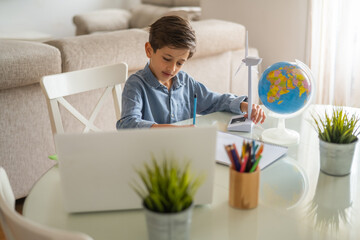 Concentrated schoolboy studying renewable energy with a wind turbine model beside a globe, taking notes and using a laptop at his desk