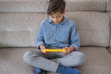 Concentrated child sitting comfortably on a sofa at home, playing video games with a bright yellow controller, fully immersed in leisure time and enjoying every moment of fun