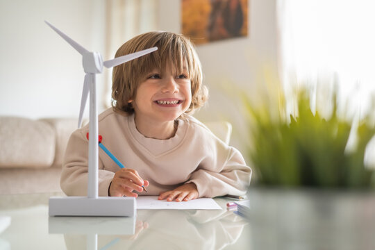 Smiling schoolboy drawing and learning about renewable energy with a small wind turbine model on a desk at home, promoting stem education and sustainability