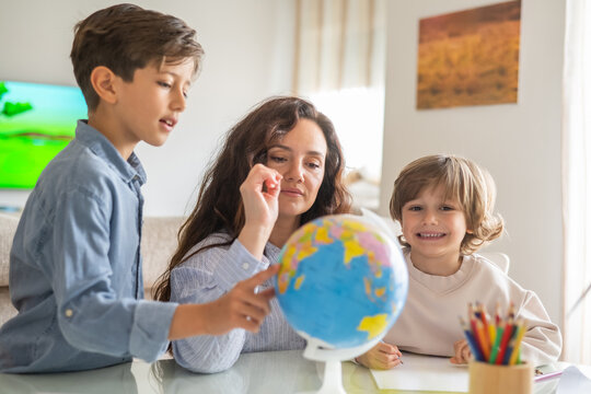 Mother pointing at globe and teaching geography to her two children during homeschooling, promoting family education and exploration of the world