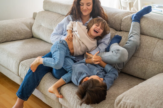 Happy family playing together on the sofa at home, mother tickling her two sons, having fun and laughing, enjoying their time together in a cozy living room