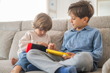 Two young brothers are sitting on a comfortable sofa in their living room, engrossed in playing...