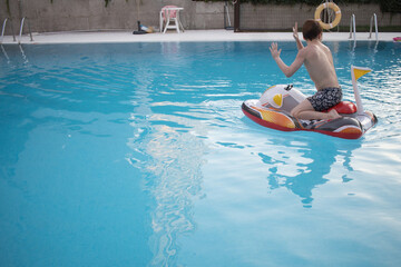 Boy floating or playing with inflatable float with motorcycle shape in the pool. Horizontal. Summer. Copy space. Faceless.