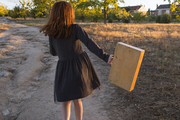 Happy readhead girl dancing and holding a wooden suitcase in a field. Horizontal, holidays