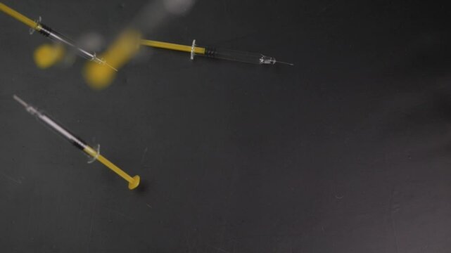 Slow-motion shot of disposable syringes dropping onto a dark matte surface, designed for vaccinations or self-administered subcutaneous injections.