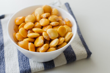 Lupin beans in a white bowl on a striped napkin on a white table. Italian food. Type of legumes. Healthy food concept. Horizontal orientation. Selective focus. Copy space.