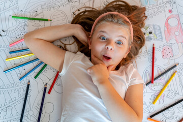 Cheerful little girl lies on the floor on a large sheet of paper with scattered colored pencils....