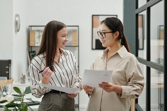 Side view portrait of two smiling young businesswomen looking at each other while discussing documents standing in modern office setting