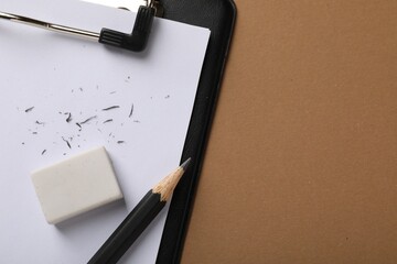 Eraser, pencil, scraps and clipboard on brown background, top view. Space for text
