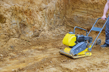 Worker operates yellow compactor to flatten soil in construction area, ensuring proper ground stability.