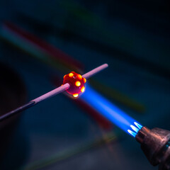 Close-up of molten glass bead being shaped with a torch flame on a rod. The glowing red bead contrasts with the dark blurred background. Concept of glassblowing craftsmanship