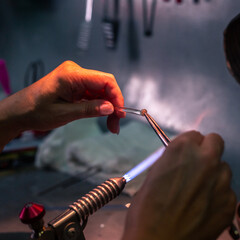 Close-up of artisan hands shaping molten glass with a blowtorch in a workshop. The flame heats the material, creating intricate designs