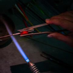 Close-up of an artisan's hands shaping molten glass using a torch flame in a workshop, with tools and materials in the background. Craftsmanship concept