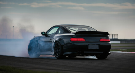 Fototapeta premium Sleek black sports car drifting on track, creating smoke and motion blur at sunset