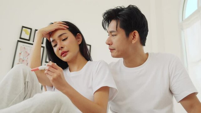 Upset Asian couple reacting to an unplanned pregnancy, woman looking distressed at a pregnancy test while man consoles her.