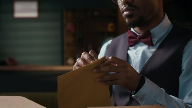 Tilt up shot of male African American writer opening envelope with letter containing good news from publishing house,sitting at table in cozy workspace