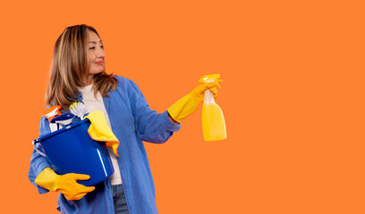 Woman prepares for cleaning with supplies in vibrant orange background, wearing gloves and holding...