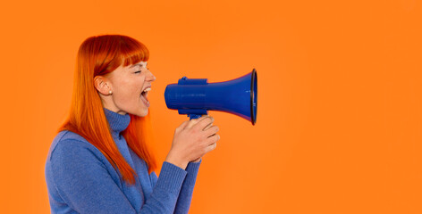 Woman with vibrant red hair passionately shouting into a blue megaphone against a bright orange background, demonstrating enthusiasm and energy in a communicative moment